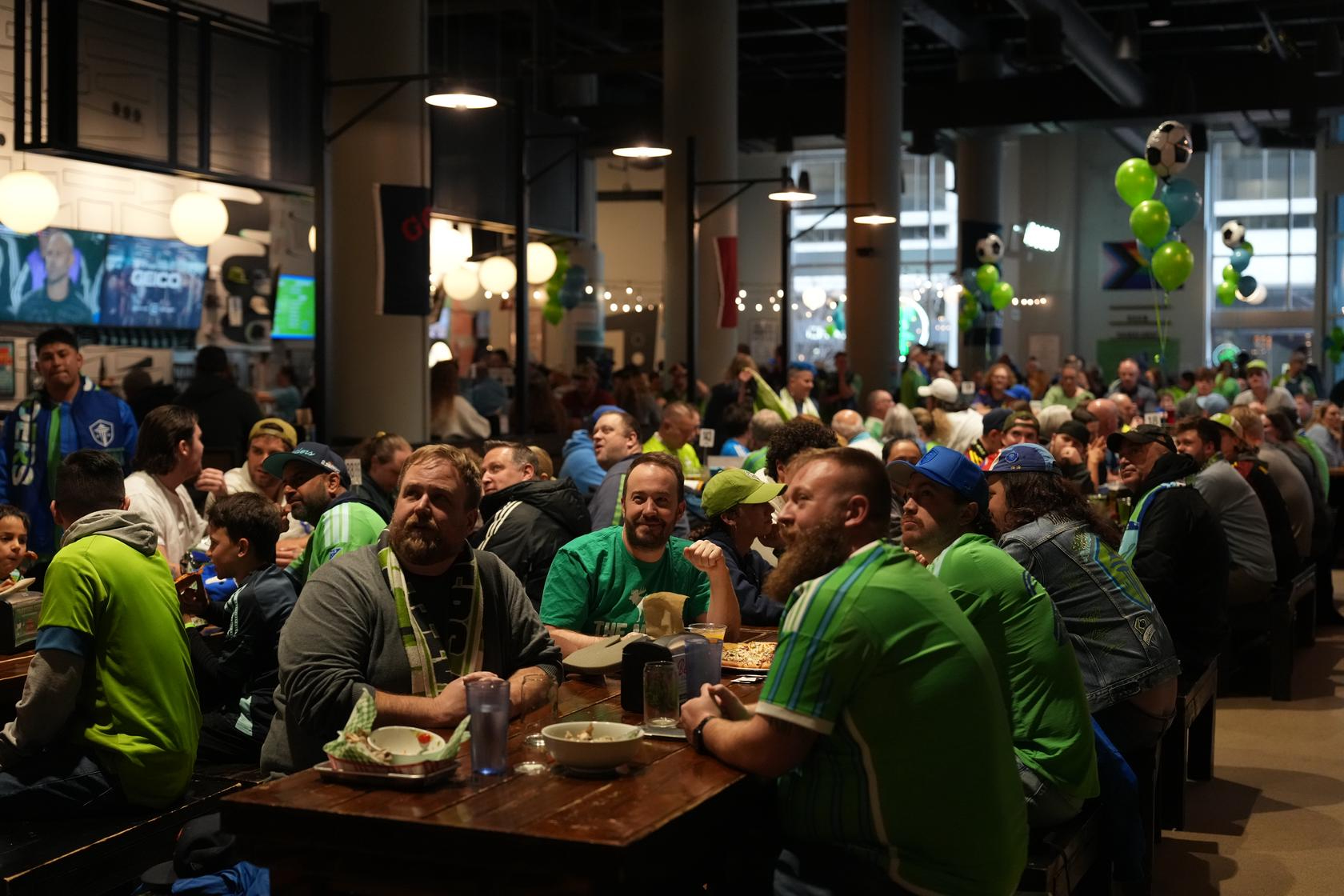 Large group of soccer fans watching a game on the big screen at Flatstick Pub around tables.