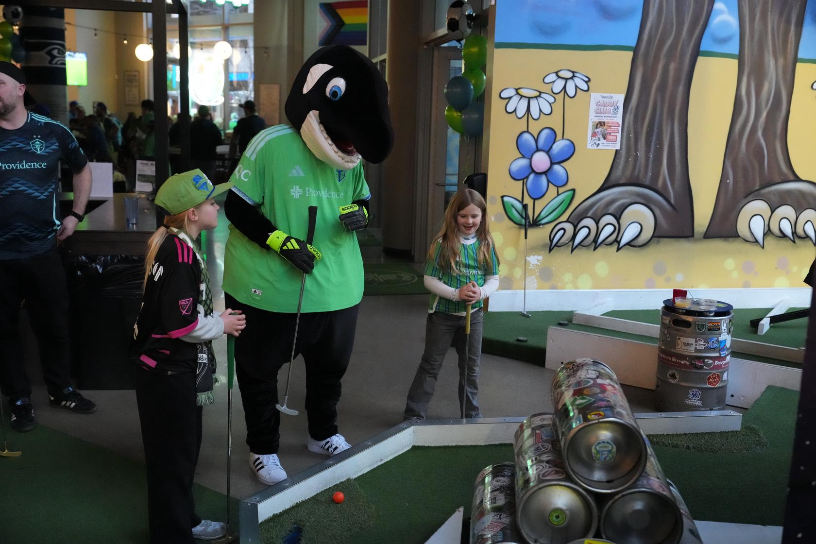 Sammy the Sounder stands around a mini golf hole with a putter in his hand, playing a game of mini golf with two young girls in their soccer jerseys at Flatstick Pub - Spokane